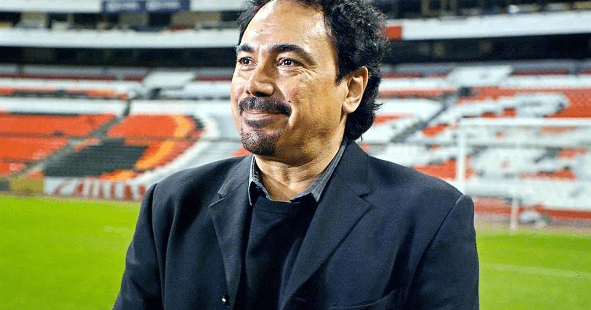 Hugo Sánchez en un estadio de fútbol, sonriendo y vestido con un traje oscuro, con gradas vacías de fondo.