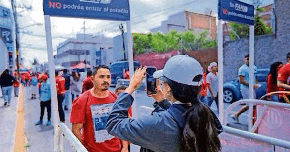 Grupo de personas con camisetas rojas esperando afuera de un estadio, con señales de restricción de entrada visibles.