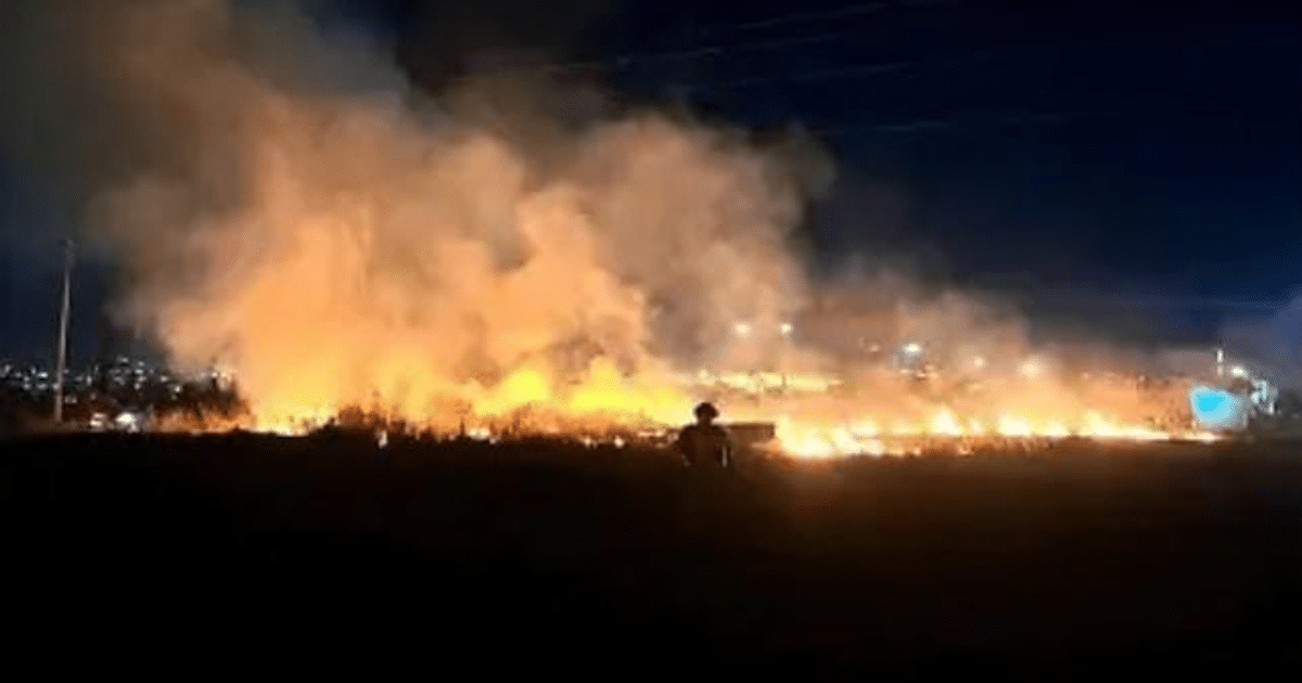 Incendio en Mazatlán con llamas y humo en un área abierta, persona observando en primer plano, luces de la ciudad al fondo.