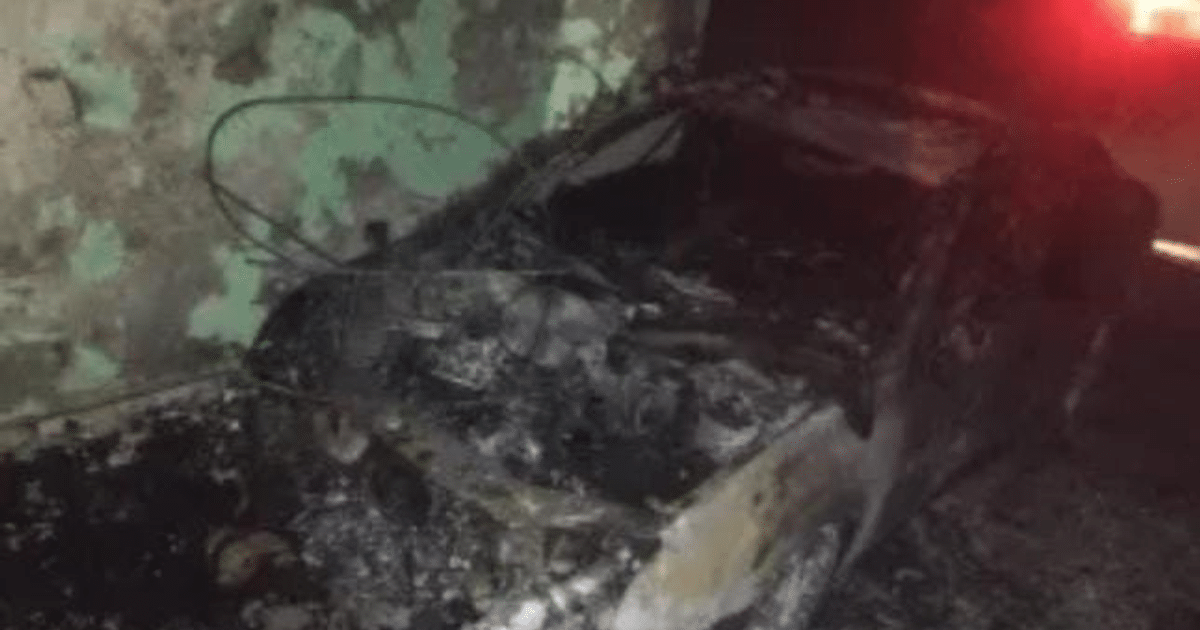 Vehículo quemado en cochera con paredes verdes descoloridas y luz roja de emergencia al fondo.