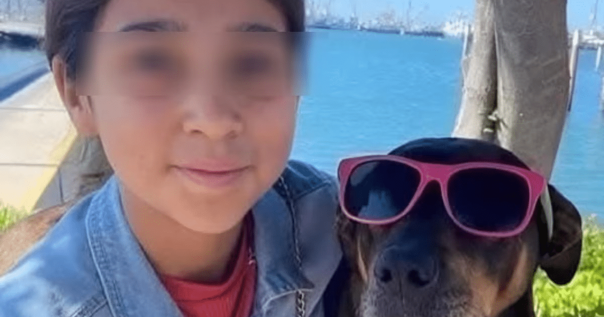 Persona sonriente con perro de gafas de sol rosas frente a un paisaje con agua y árboles.