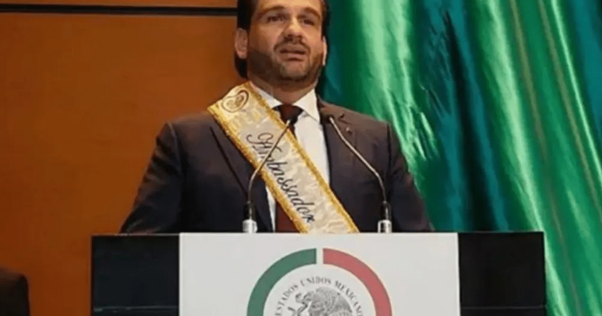 Hombre en podio con banda de 'Embajador' en evento oficial, cortina verde y emblema mexicano.