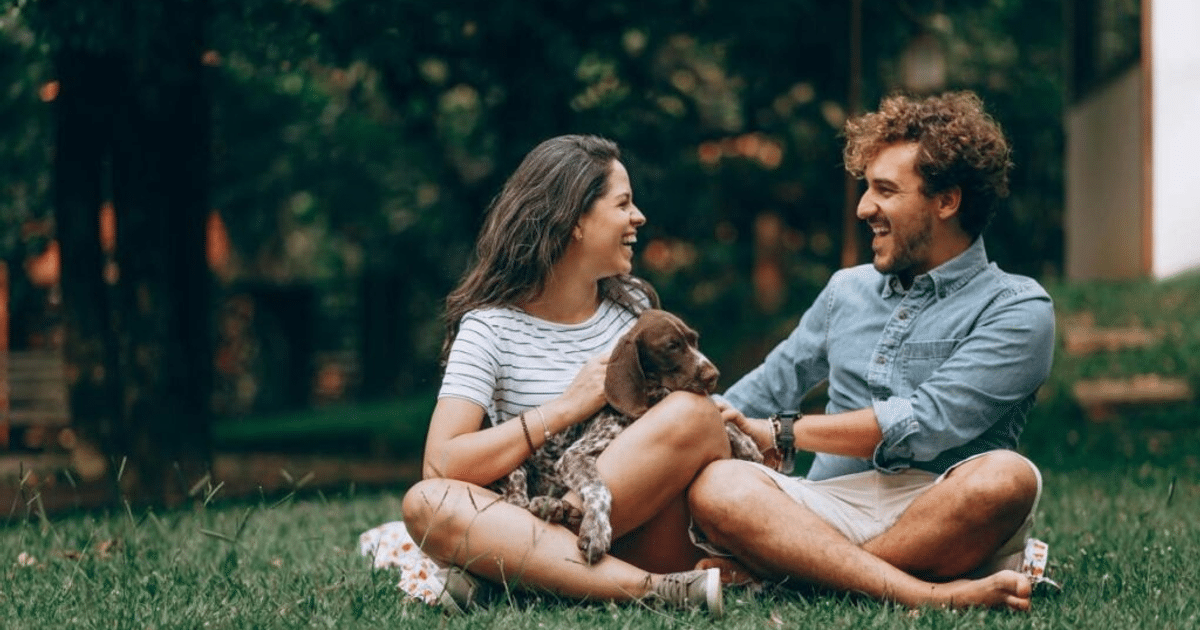 Pareja sentada en el césped con un perro, disfrutando de un día soleado.