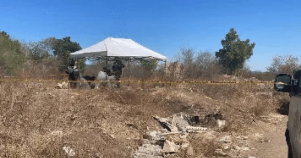 Investigadores bajo un toldo blanco en una narcofosa en Culiacán, rodeados de cinta amarilla y escombros en un entorno desértico.