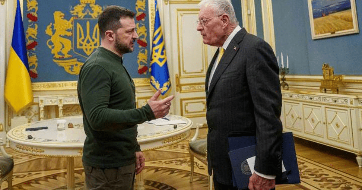 Dos hombres en una reunión formal discutiendo sobre el conflicto ucraniano, uno con suéter verde y otro con traje gris, en un entorno decorado con banderas.