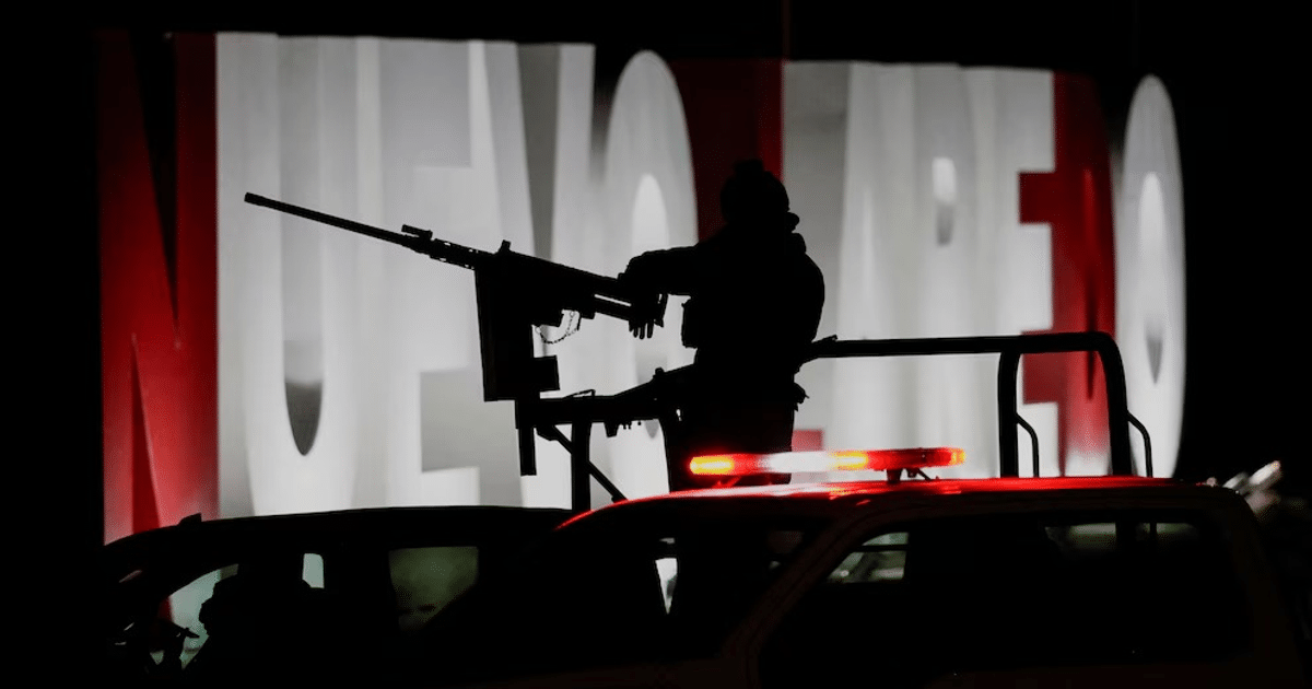 Hombre armado en vehículo con letrero de Nuevo Laredo al fondo, escena nocturna de seguridad.