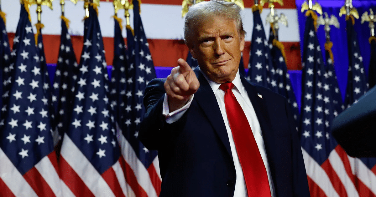 Funcionario estadounidense en traje oscuro y corbata roja, con banderas de EE.UU. al fondo, anunciando cambios en política de seguridad.