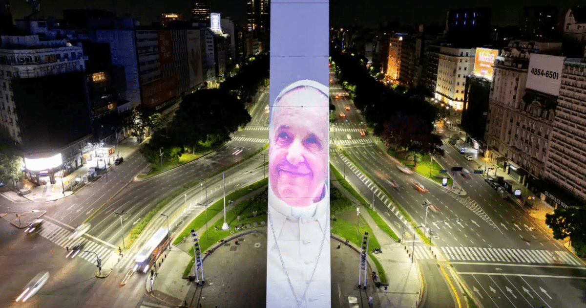Obelisco de Buenos Aires iluminado con la imagen del Papa Francisco, rodeado de tráfico y edificios iluminados.