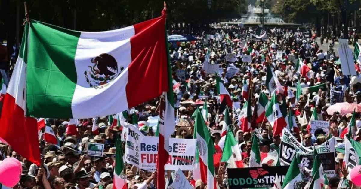 Multitud en manifestación con banderas de México y carteles de protesta en una calle amplia.