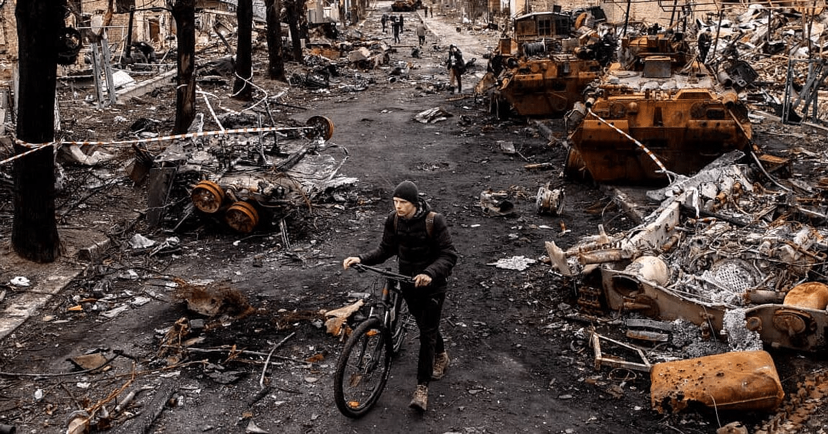 Hombre empujando bicicleta en calle devastada por conflicto, rodeado de escombros y vehículos destruidos.