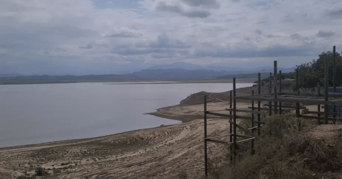 Paisaje de presa en Sinaloa con cuerpo de agua, tierras áridas y montañas al fondo, bajo un cielo nublado.