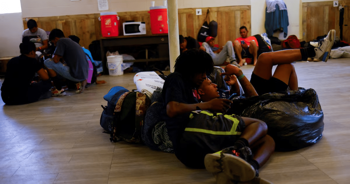 Personas en un refugio para migrantes en Texas, algunas sentadas y otras en el suelo, en un ambiente de acogida con paredes de madera.