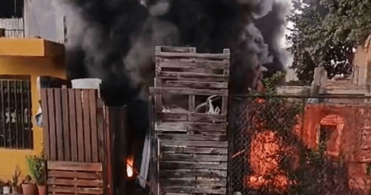 Incendio en Santa Elena con humo negro y llamas en una estructura de madera, edificio amarillo al fondo.