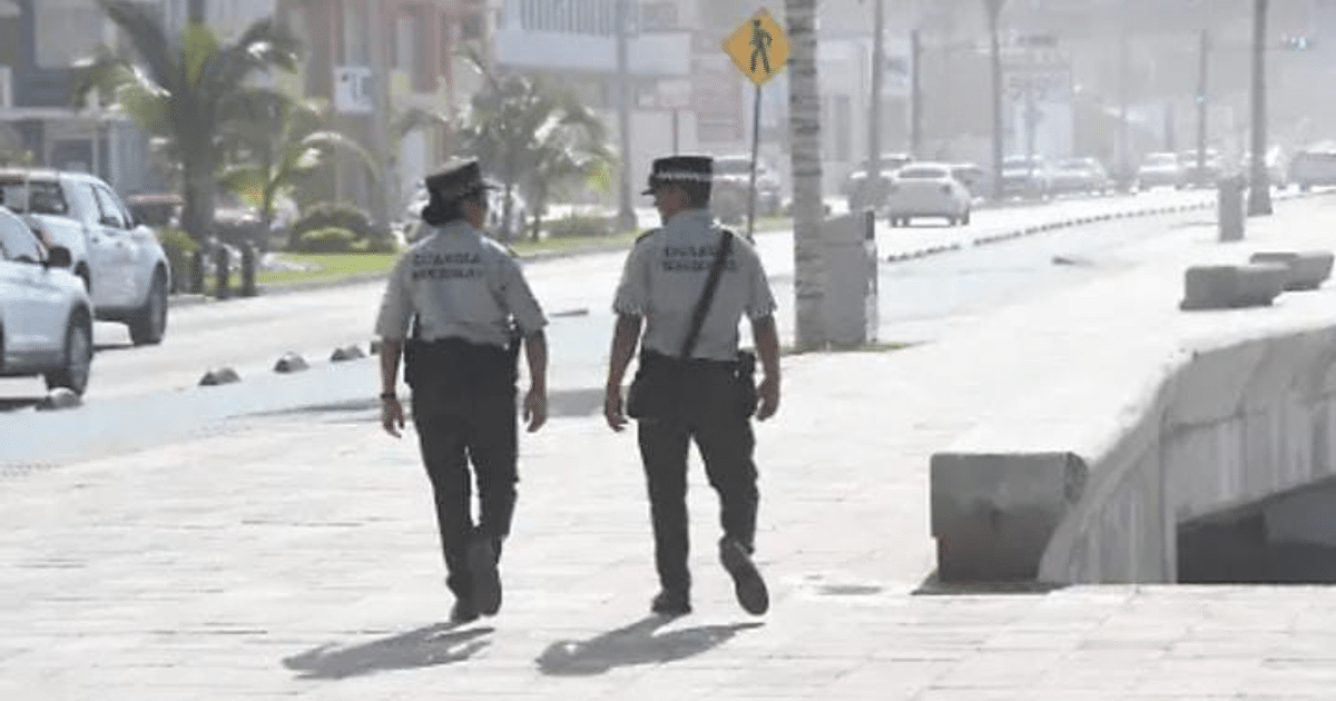 Dos oficiales de policía caminando en una acera con vehículos y palmeras al fondo, en un operativo de seguridad en Mazatlán.