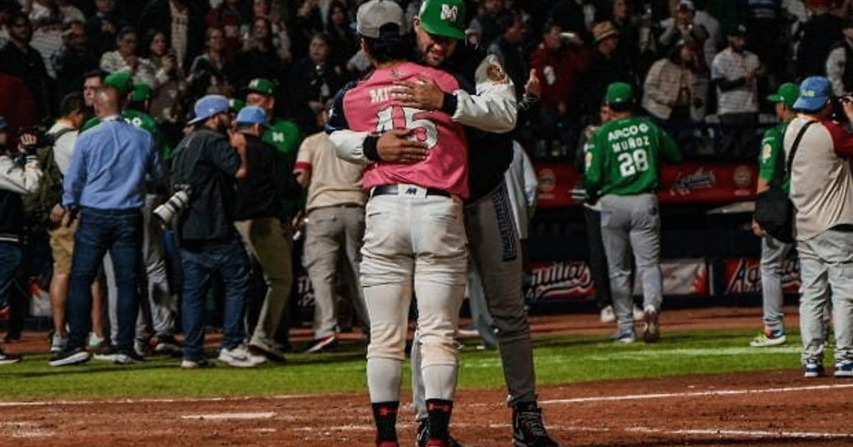 Dos personas abrazándose en un campo de béisbol, una con camiseta rosa número 15, rodeadas de jugadores y fotógrafos, celebrando una victoria.