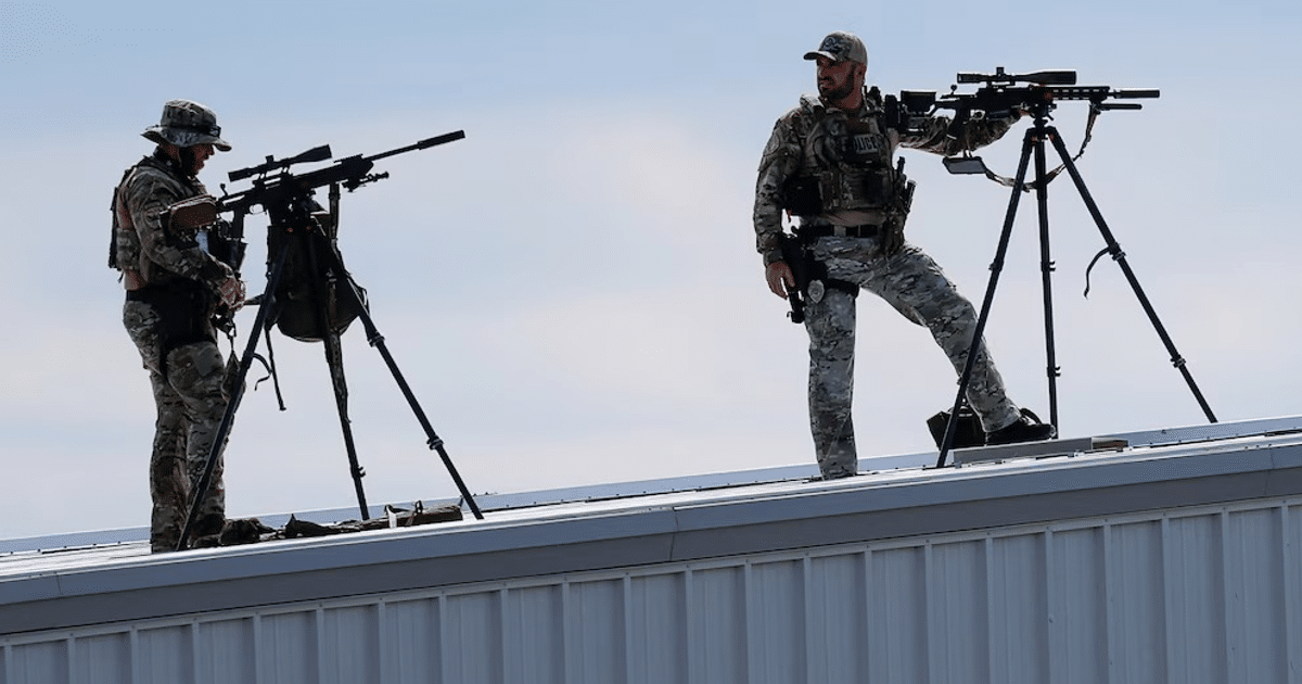 Dos francotiradores en un techo con rifles montados en trípodes, cielo despejado de fondo.