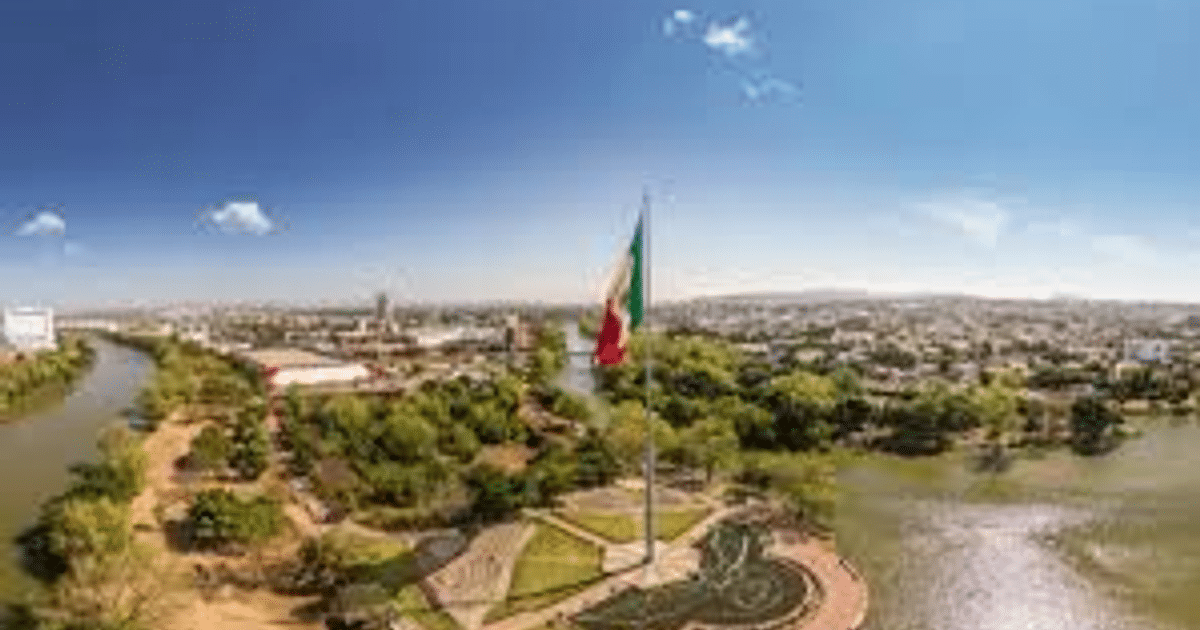 Vista panorámica de un paisaje urbano en Sinaloa con un río, áreas verdes y la bandera de México ondeando.