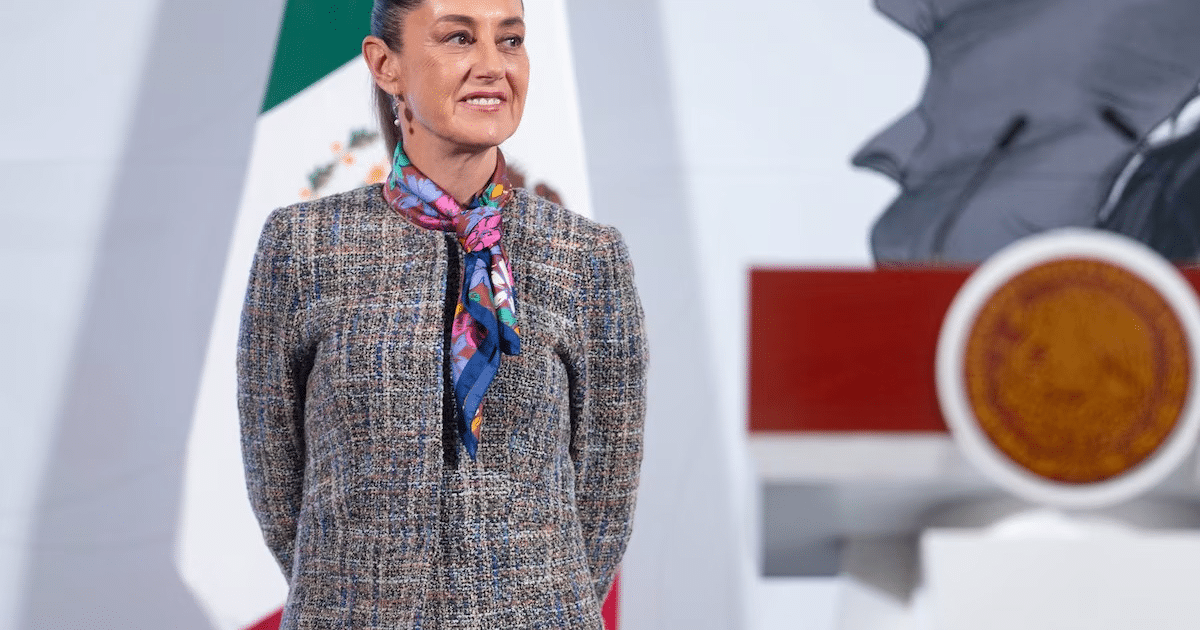 Mujer sonriente con abrigo claro y bufanda colorida frente a la bandera de México en un evento formal.