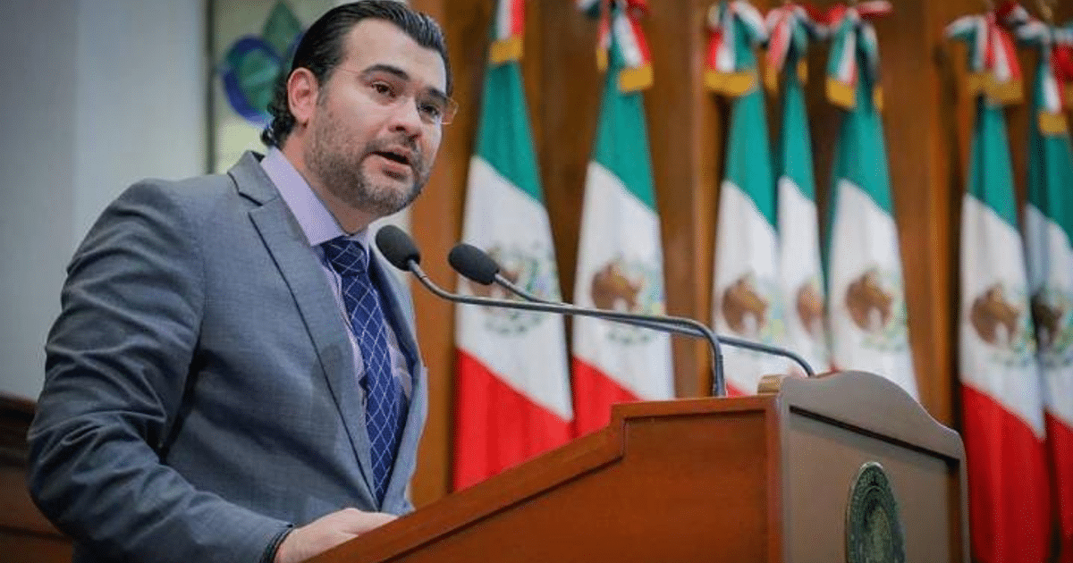 Hombre en traje gris hablando en un podio con banderas de México detrás