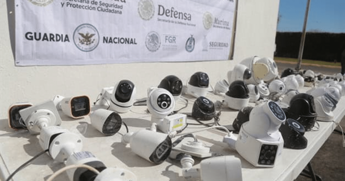 Mesa con cámaras de vigilancia de diferentes tipos y colores, con una pancarta de seguridad al fondo.