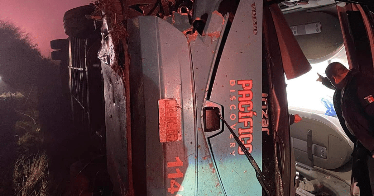 Camión volcado con logotipo 'PACÍFICO DISCOVERY' en un área rural, iluminado por luces de emergencia, con dos personas inspeccionando.