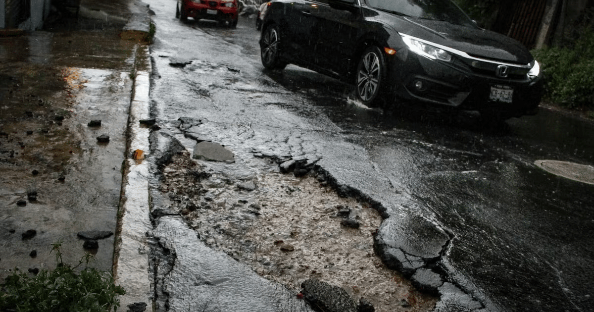 Calle lluviosa con bache y autos circulando, reflejando condiciones adversas de las vías urbanas.