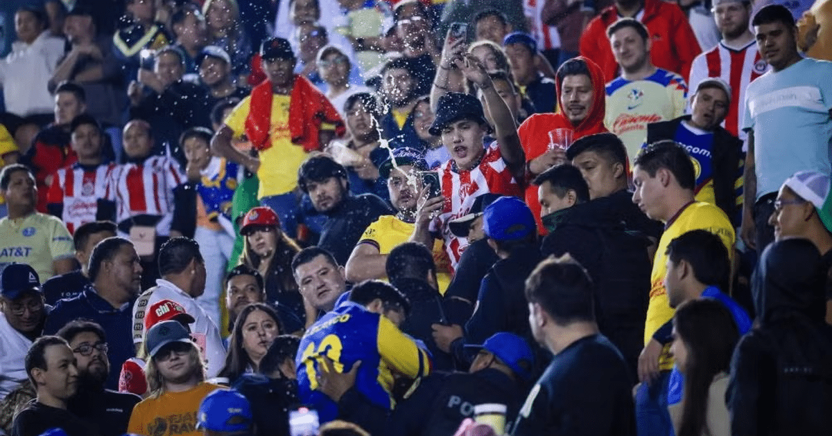 Aficionados en un evento deportivo, algunos celebrando y otros serios, con agua en el aire, durante el Clásico Nacional entre América y Chivas.