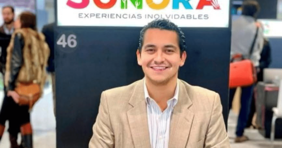 Hombre sonriendo frente a cartel de Sonora, con saco claro y camisa de rayas, en un evento promocional.
