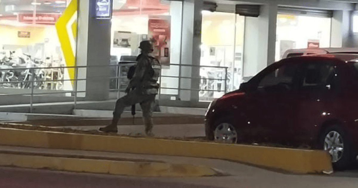 Militar armado de pie en un entorno urbano nocturno en Culiacán, cerca de un edificio comercial y un automóvil rojo.