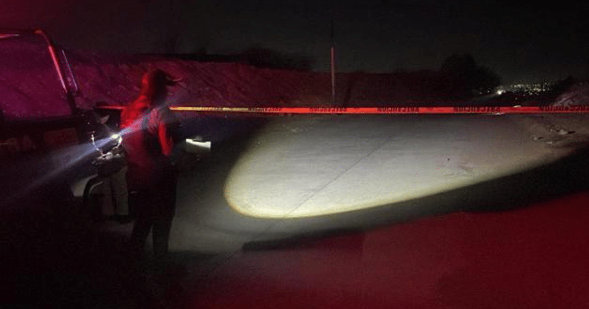 Persona con linterna en escena nocturna de investigación policial, área delimitada por cinta de advertencia.