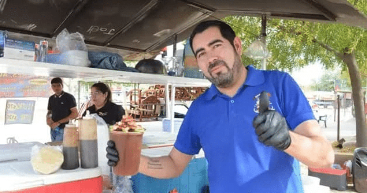 Hombre con vaso de comida en feria al aire libre, guantes negros, camiseta azul, salsas e ingredientes en el fondo.