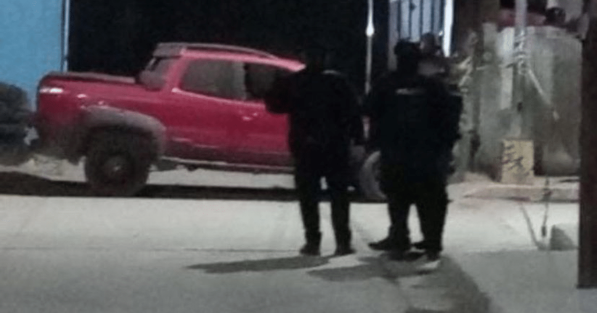 Automóvil rojo estacionado de noche con dos personas vestidas de negro cerca, en un entorno tranquilo.