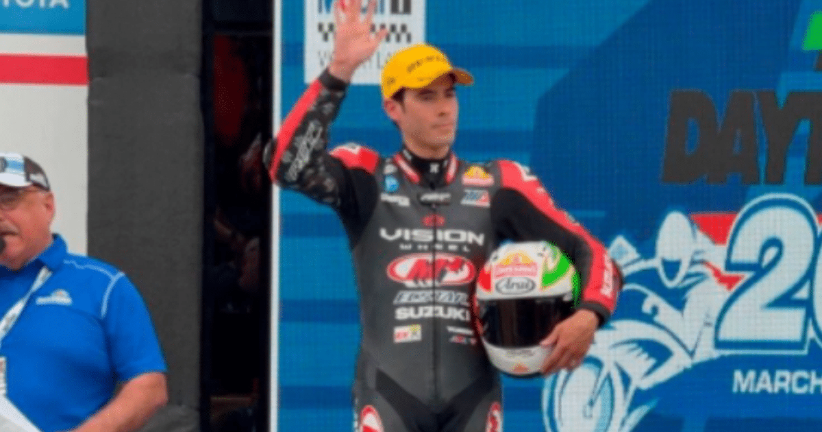 Piloto de motociclismo saludando en Daytona con casco en mano, junto a un hombre con camiseta azul.