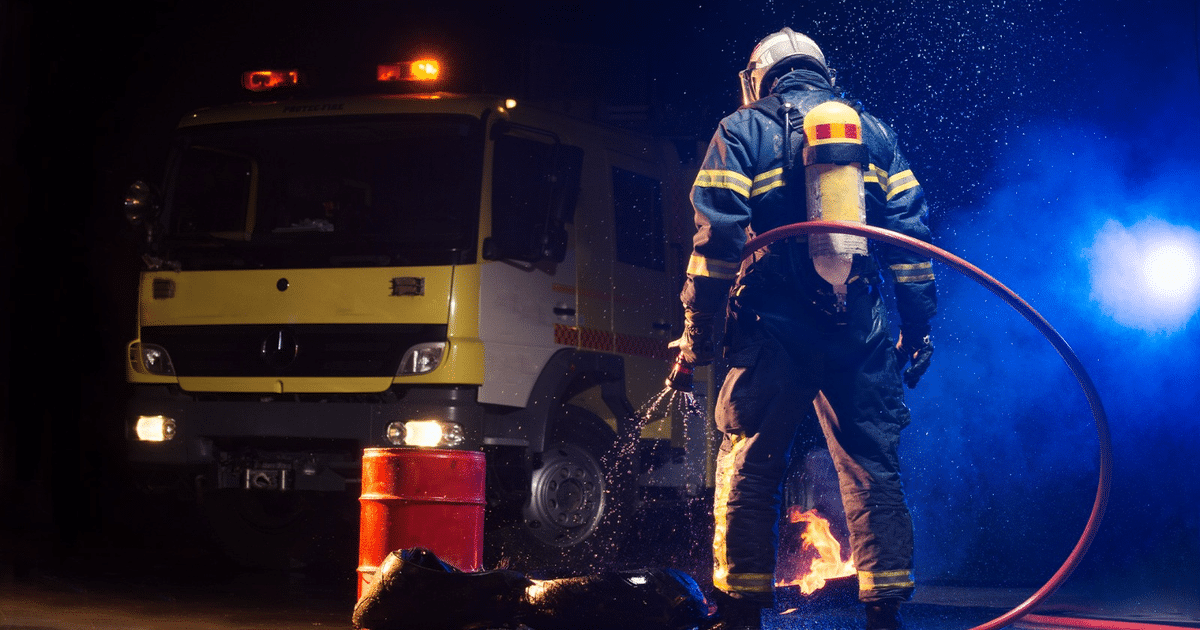 Bombero apagando fuego con manguera, barril rojo y camioneta de bomberos amarilla en el fondo.