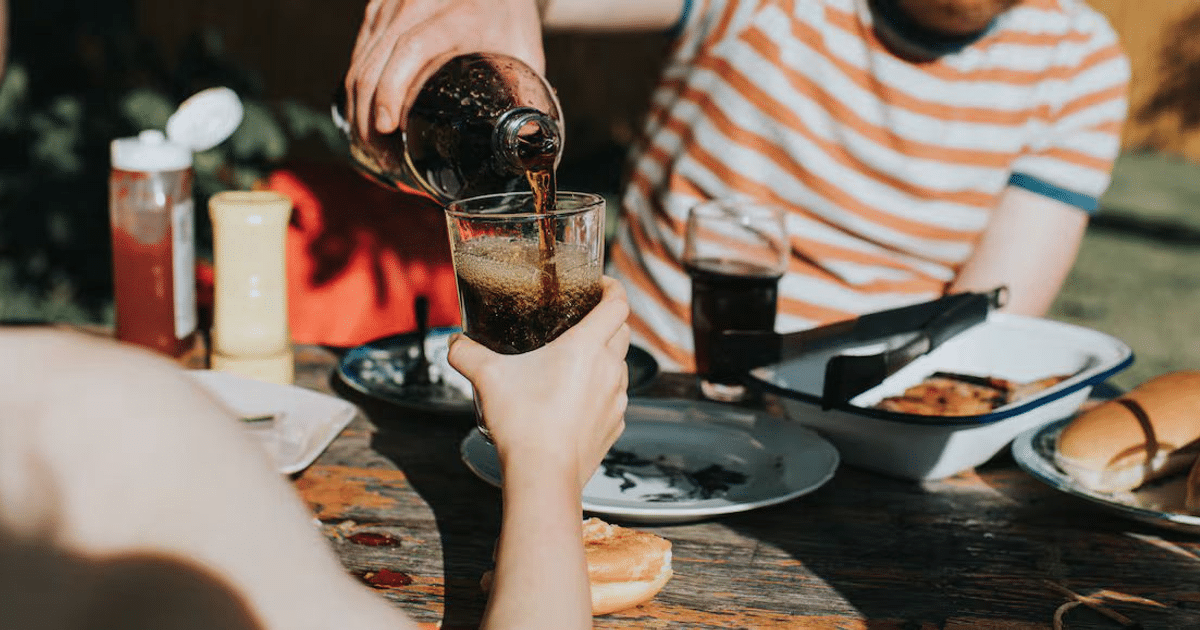 Persona sirviendo bebida oscura en vaso durante comida al aire libre, con platos de comida en mesa de madera.