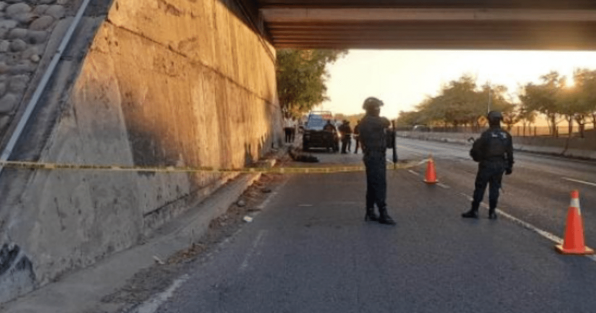 Policías armados aseguran una carretera en Culiacán tras el hallazgo de un cuerpo envuelto en plástico, con un vehículo policial al fondo y la luz del atardecer iluminando la escena.