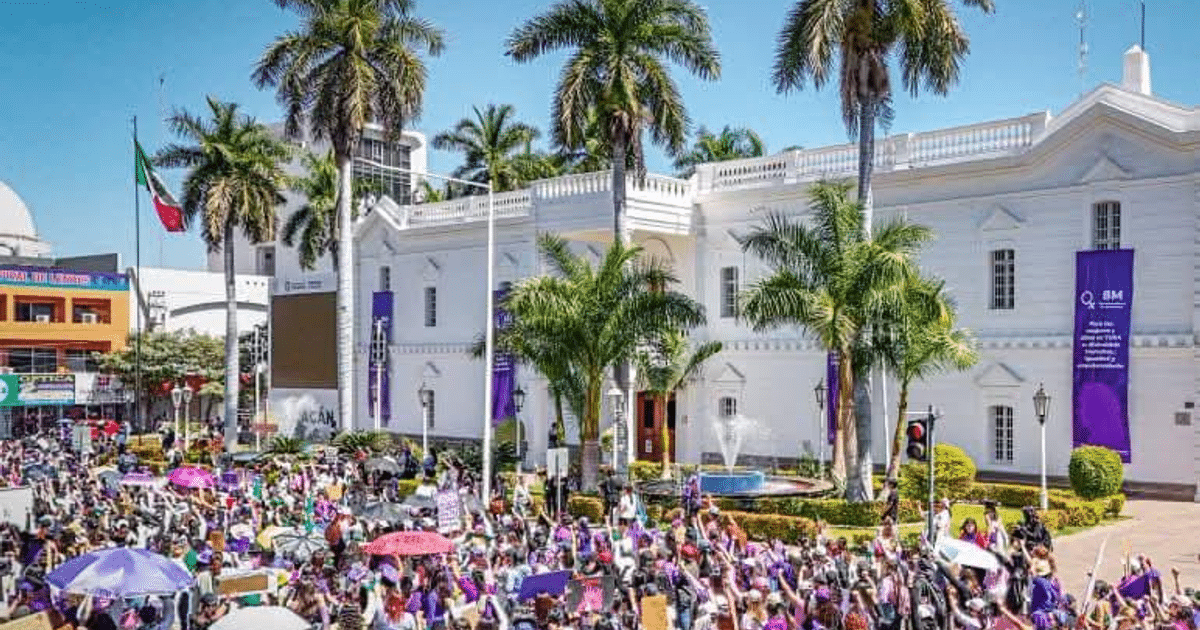 Manifestación en Sinaloa por el Día Internacional de la Mujer, con personas portando pancartas y sombrillas coloridas frente a un edificio gubernamental.