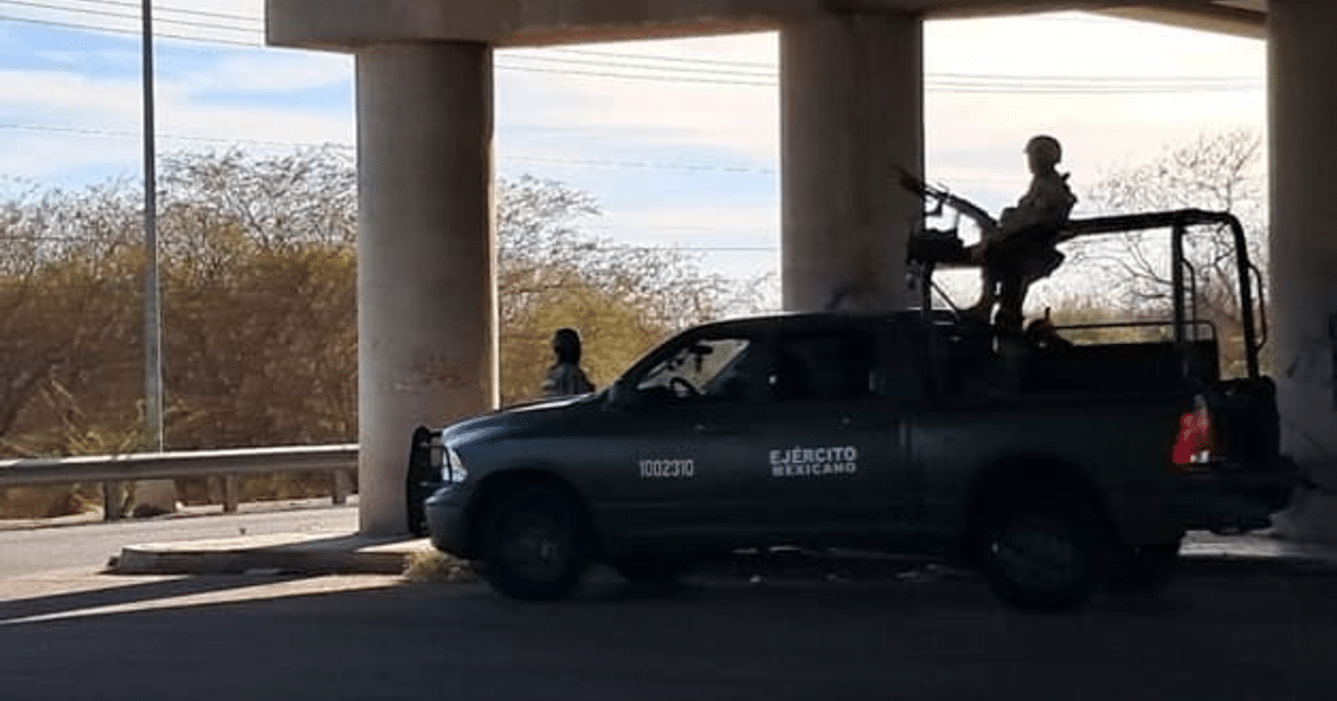 Vehículo militar del ejército mexicano bajo un puente con soldado armado y persona cercana, en un paisaje árido.