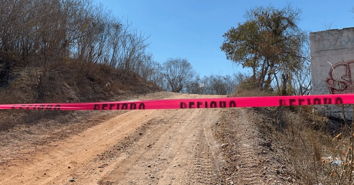 Camino de tierra bloqueado por cinta de peligro rosa en Mazatlán, Sinaloa, con vegetación seca y estructura con grafitis.