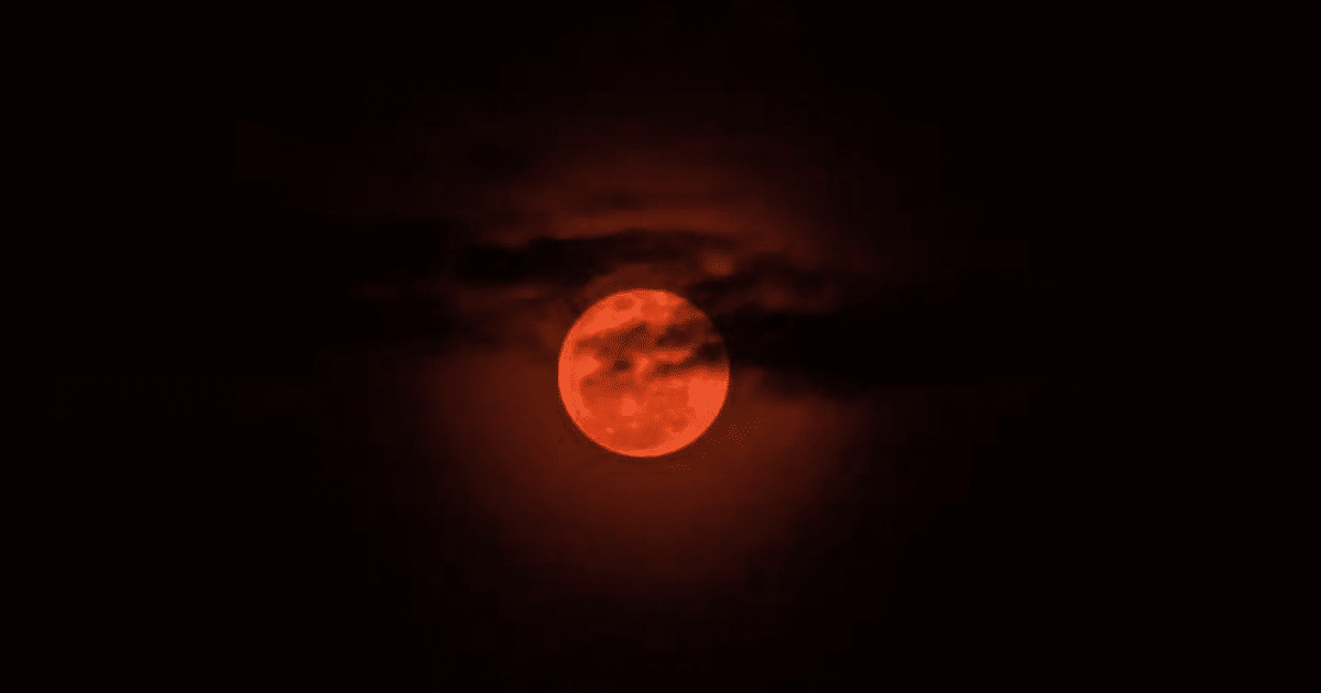 Imagen de una luna llena roja durante un eclipse lunar, parcialmente cubierta por nubes en un cielo oscuro.