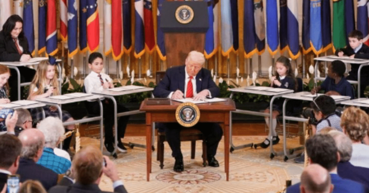 Niños en evento educativo formal con hombre escribiendo en escritorio, relacionado con políticas educativas de Trump.