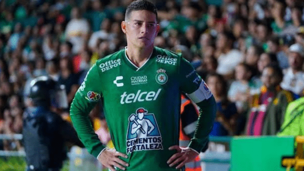 Jugador de fútbol con uniforme verde en partido, espectadores al fondo, oficial de seguridad a la izquierda.
