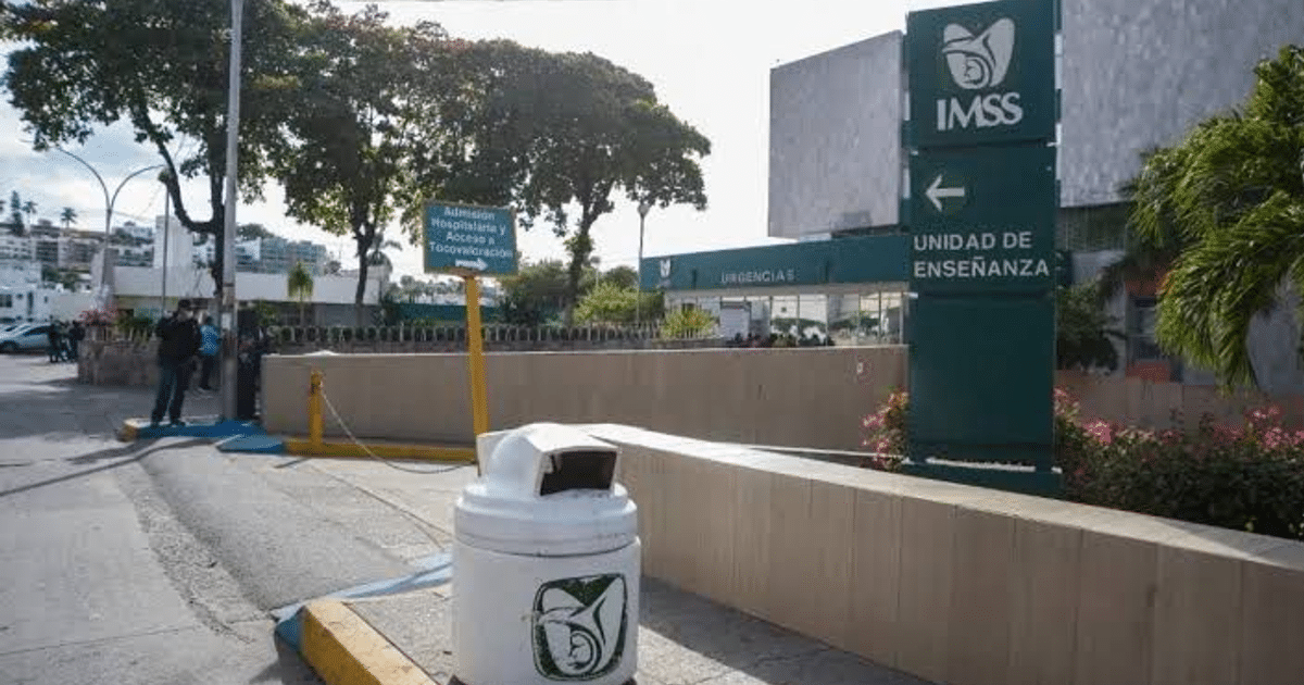 Entrada de hospital con carteles de admisión y unidad de enseñanza, basurero en primer plano, personas y vegetación al fondo.