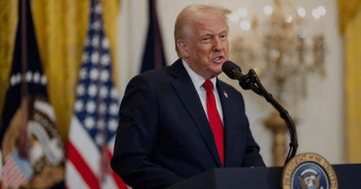 Persona hablando en un micrófono durante una conferencia sobre la fusión de la DEA y ATF, con banderas y fondo decorativo.