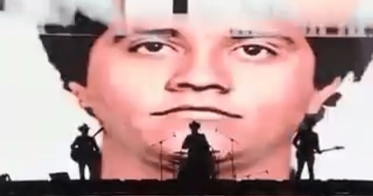 Proyección de rostro destacado en fondo durante concierto con músicos en silueta en escenario, representación de homenaje a líder criminal.