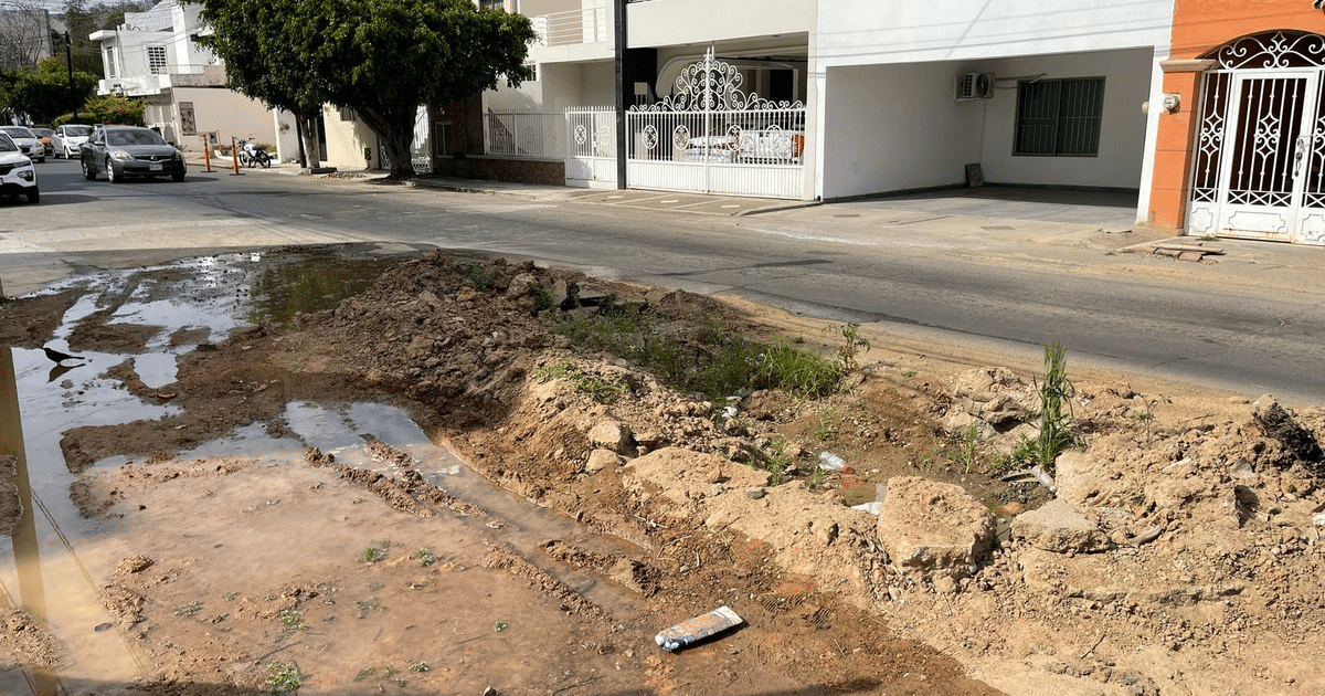 Socavón en la calle Río Baluarte con tierra removida y agua estancada, rodeado de basura y vegetación, con vehículos y edificios al fondo.