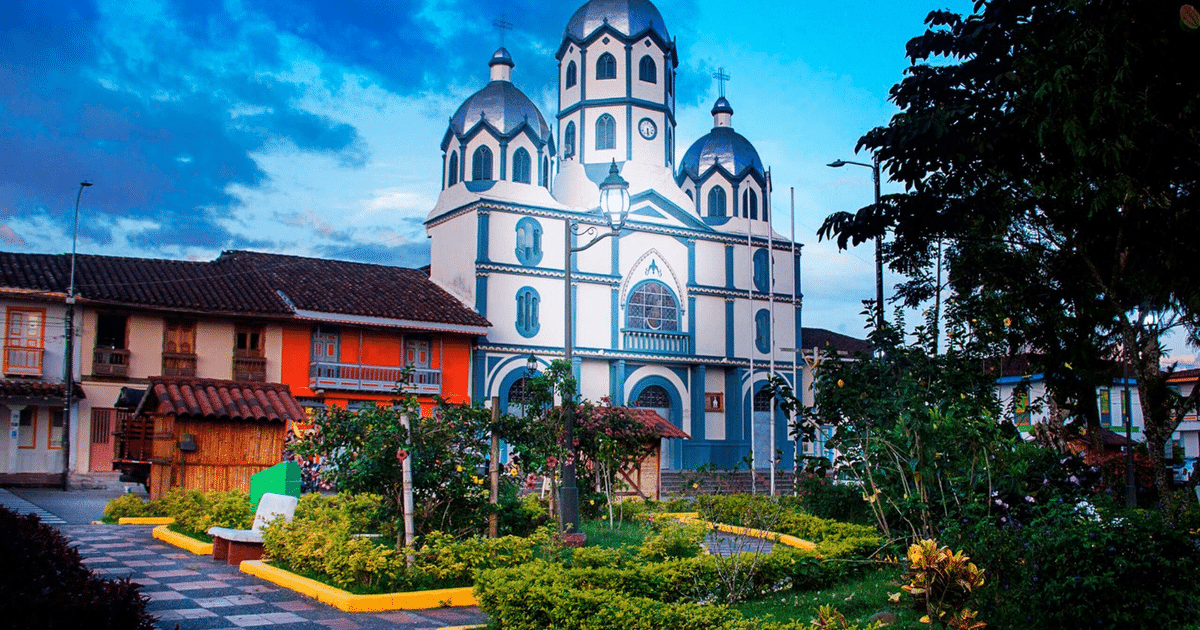 Iglesia con cúpulas blancas y azules en un jardín florido, rodeada de casas coloridas bajo un cielo azul.