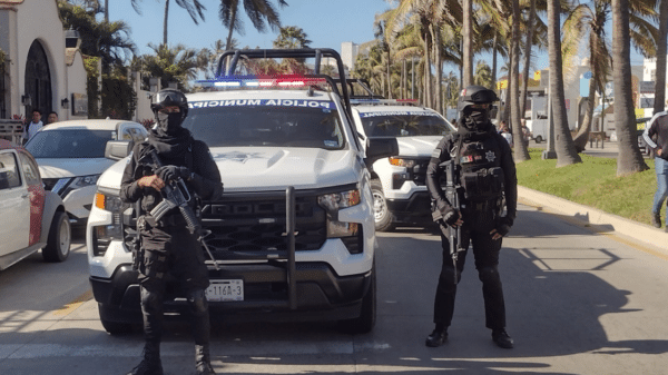 Dos policías armados frente a un vehículo policial en una calle urbana con palmeras, en Mazatlán.
