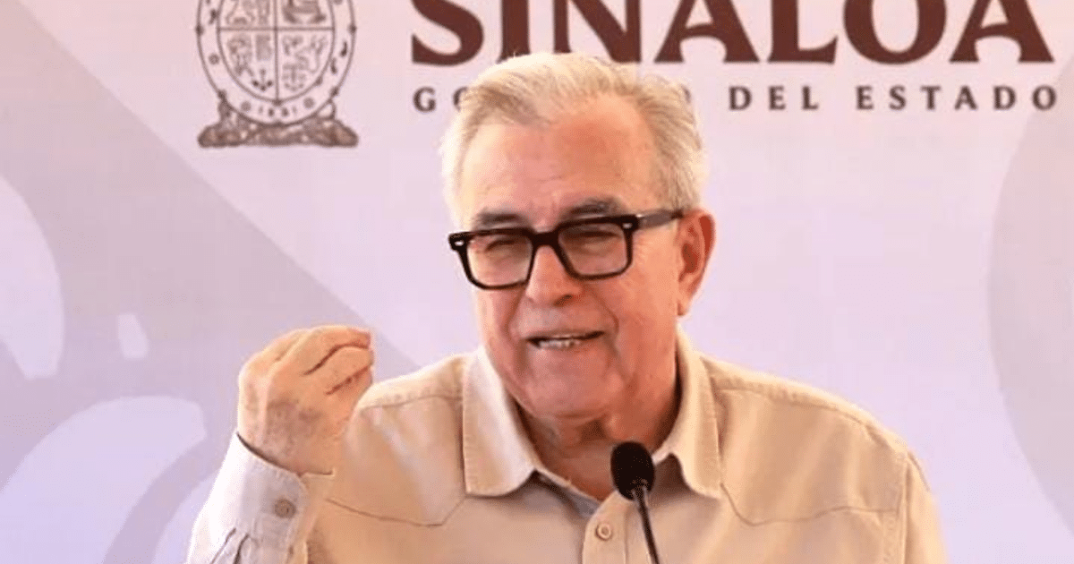 Persona hablando en un evento gubernamental en Sinaloa con el logo del estado de fondo.