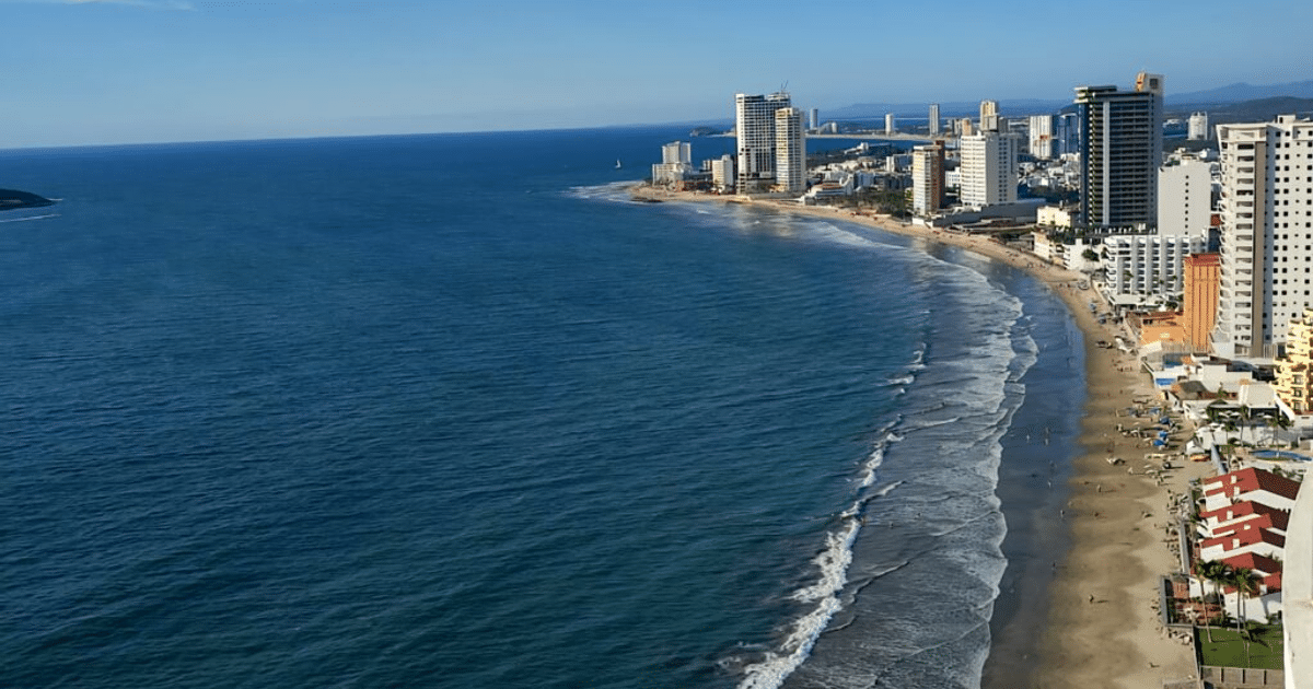Playa de Mazatlán con aguas azules, edificios altos y un muelle, bajo un cielo despejado.
